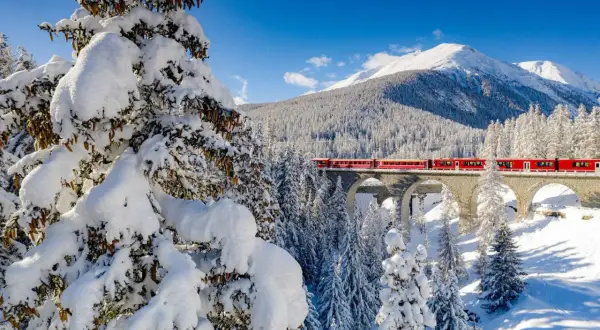 Röda tåget korsar en snöig bro i Schweiz, ett drömresmål med majestätiska berg och vinterlandskap.