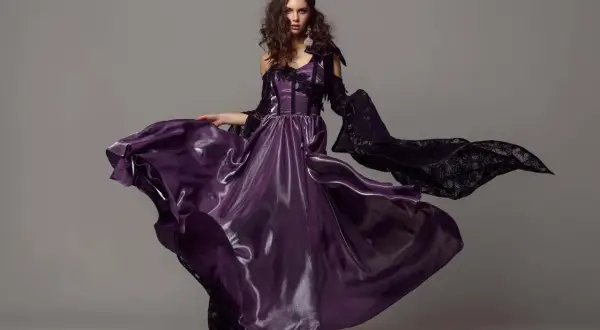 A model in a flowing purple gown and black lace sleeves poses for studio fashion photography.