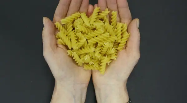 Dłonie trzymające porcję żółtego makaronu fusilli na ciemnym tle.