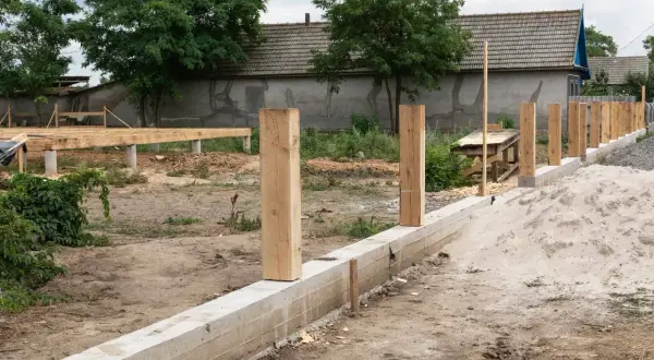 Solidny fundament pod ogrodzenie z betonowym murem i drewnianymi słupkami, w tle budowa domu.