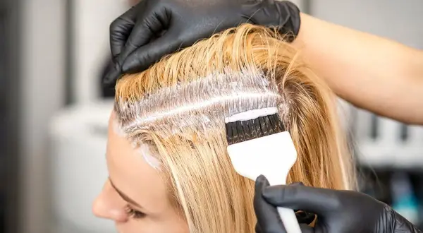 Fryzjerka w czarnych rękawiczkach nakłada farbę na blond włosy, wykonując farbowanie odrostów.