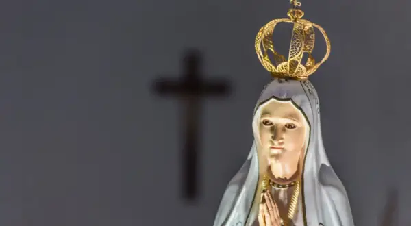Figurka Matki Bożej z Fatimy, w koronie, z modlącymi się dłońmi. W tle niewyraźny krzyż, symbol objawienia fatimskiego.