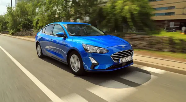Błękitny Ford Focus sedan pędzi po miejskiej drodze, mijając drzewa i budynki.