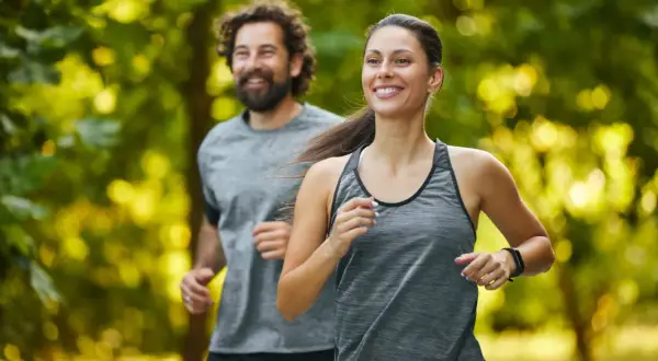 Coppia sorridente corre nel parco. Lei, in primo piano, è determinata, lui la segue con gioia. Un esempio di come iniziare a correre a 50 anni sia possibile e appagante.