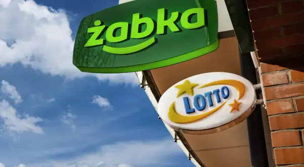 Zielona tablica "Żabka" i biała z napisem "LOTTO" wiszą na tle błękitnego nieba z chmurami.