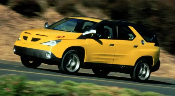 Żółty Pontiac Aztek jedzie po drodze, otoczony rozmytą zielenią i brązem.