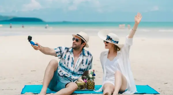 Para na plaży robi selfie z kamerką GoPro, ciesząc się wakacjami. W tle błękitne morze i niebo.