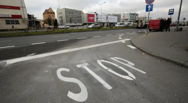 Na asfalcie widoczna jest biała linia warunkowego zatrzymania "STOP". W tle budynki i ruch uliczny.
