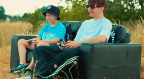 Dwóch chłopców gra w gry wideo na czarnej kanapie na łonie natury. Jeden z nich, młodszy, ma na głowie czarną czapkę typu bucket hat, a drugi, starszy, nosi okulary przeciwsłoneczne. Wyglądają jakby byli na planie teledysku rapera.