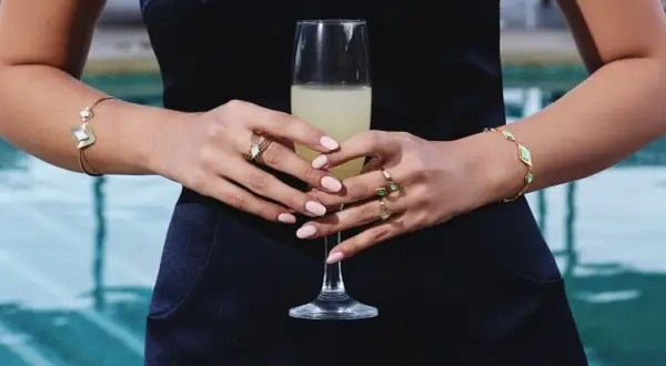 Manos con uñas rosadas y varios anillos dorados, sosteniendo una copa de champán. Brazaletes dorados adornan ambas muñecas.