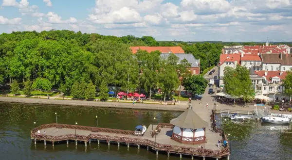 Drewniany pomost z altaną nad jeziorem, zielone drzewa i miasteczko z czerwonymi dachami. Idealne miejsce, gdzie na Mazury.