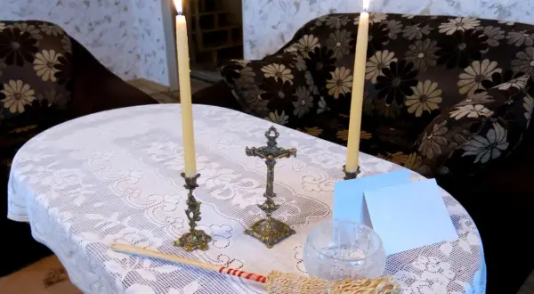 Dwa płonące świeczniki, krzyż, kropidło i białe kartki na stole przykrytym koronkowym obrusem.