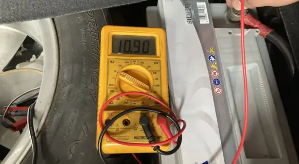 Multímetro amarillo muestra 10.90V al comprobar bateria coche. Se ve la batería, un neumático y cables.