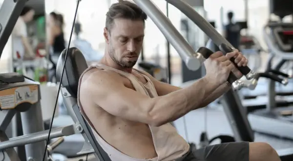 Mann trainiert im Fitnessstudio, um schnell zuzunehmen. Er sitzt auf einem Gerät und drückt die Griffe nach vorne.