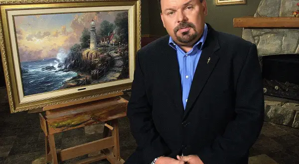 A man in a suit sits beside a Thomas Kinkade painting of a lighthouse on a cliff.