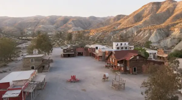 Un pueblo del oeste con edificios de madera y un pozo de agua, rodeado de colinas áridas.