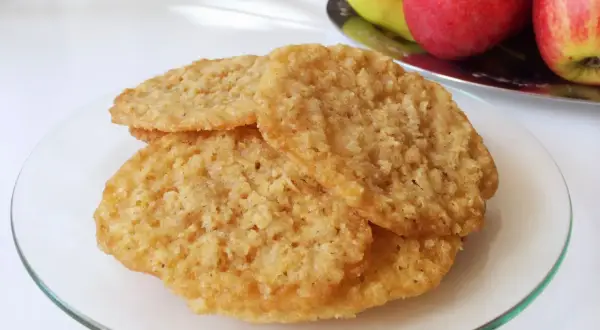 Tre gyllene glutenfria havreflarn ligger staplade på en glasfat. I bakgrunden syns äpplen och päron.