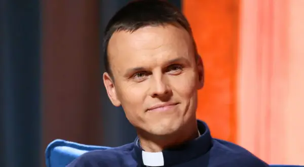 Uśmiechnięty ksiądz z osiedla, młody duchowny w sutannie, z uwagą słucha rozmówcy.