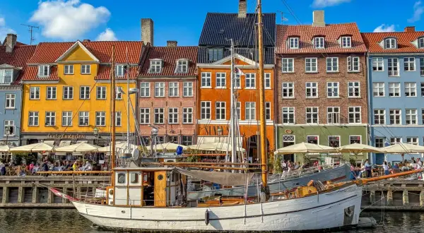 Ein weißes Segelschiff liegt im Hafen vor bunten Häusern. Menschen sitzen in Cafés am Wasser. Ein perfekter Tag im kopenhagen sommer.