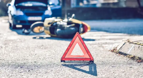 Trójkąt ostrzegawczy na drodze po wypadku. Złoty skuter leży na boku, obok niebieski samochód z otwartą maską. Może to być symbol problemów z ubezpieczeniem, jak 2 rata OC po terminie.