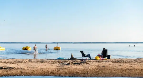 Solig dag vid Karlstad Swecamp Bomstadbaden. Folk badar och kopplar av på stranden med gula trampbåtar i vattnet.