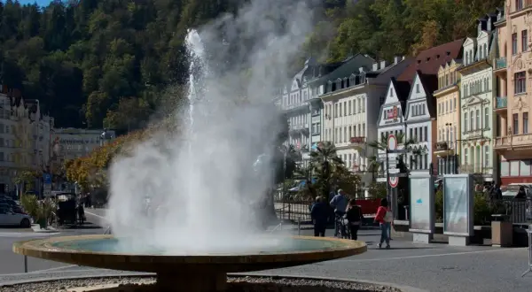 Der sprühende geysir karlsbad mit dampfender Fontäne vor bunten Häusern und grünen Bäumen.