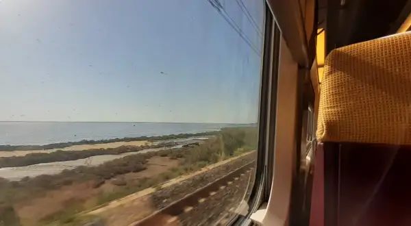 Blick aus dem Zugfenster auf eine Küstenlandschaft mit Meer und Bahngleisen. Die Reise mit dem Zug nach Mallorca beginnt.