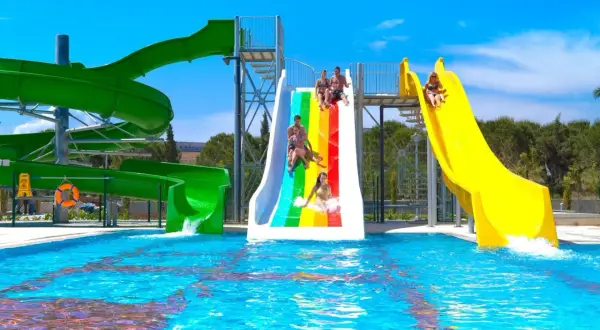 Wesoła zabawa na zjeżdżalniach wodnych w Venosa Beach Resort & Spa. Dzieci i dorośli ścigają się po tęczowej i żółtej zjeżdżalni, wpadając do basenu.