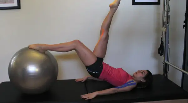 Donna con taping blu e arancione esegue un esercizio di pilates con palla, attivando i muscoli paravertebrali.