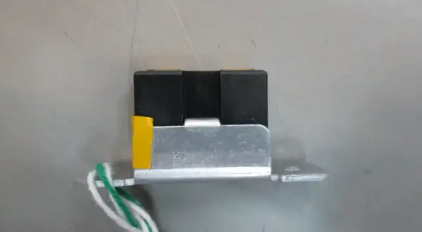 Mały, czarny element z żółtym akcentem i przewodami, przypominający wtyczkę RGB, zamontowany na metalowej płytce.