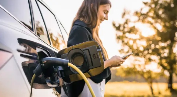 En kvinna laddar sin elbil utomhus, med solen som lyser genom träden. En gul laddningskabel är ansluten till bilen.
