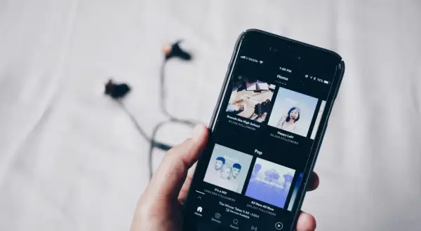 Dłoń trzyma smartfon z aplikacją do streaming muzyki. W tle leżą słuchawki.