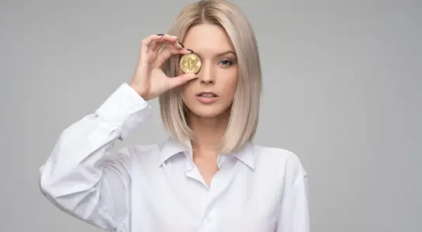 Blondynka w białej koszuli trzyma monetę bitcoin przed okiem, czując zazdrość, że ktoś ma lepiej.