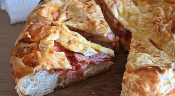 Una torta rustica con pasta sfoglia ripiena di salumi e pomodoro, tagliata a fette su un tagliere di legno.