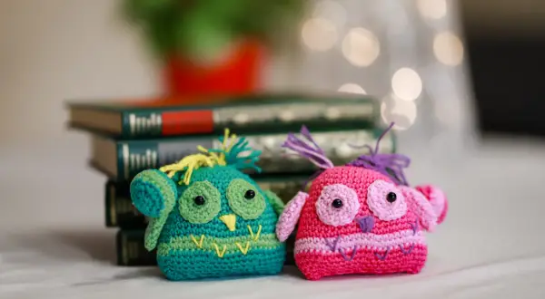 Due gufi all'uncinetto, uno verde e uno rosa, seduti davanti a libri antichi. Un dolce schema per creare questi simpatici amici.