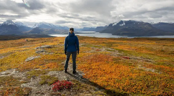Une randonneuse admire un paysage automnal aux couleurs vives, parfait pour randonner en novembre en France. Montagnes enneigées et lac en arrière-plan.