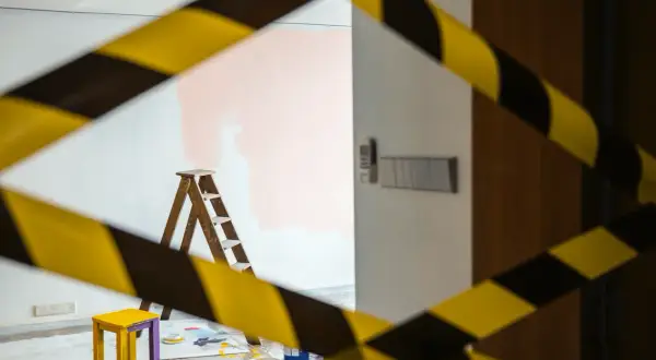 Cinta de precaución amarilla y negra rodea una habitación con una escalera, un taburete y pintura rosa en la pared, ilustrando la diferencia entre obras de mejora y obras de conservación.