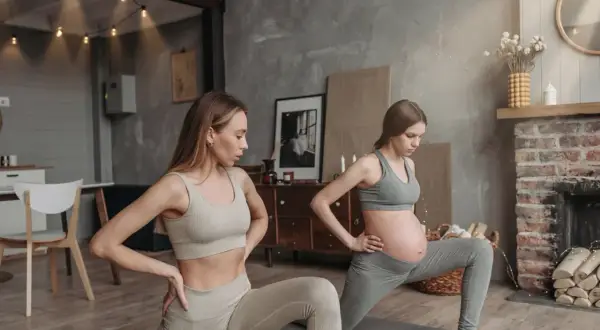 Två kvinnor gör yoga gravid i ett mysigt rum. Den ena kvinnan är gravid och den andra är hennes träningskompis.