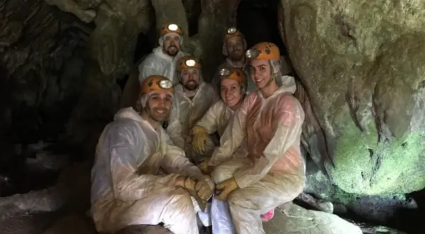 Grupo de exploradores sonríe dentro de la Cova do Rei Centolo, equipados con cascos y trajes protectores.