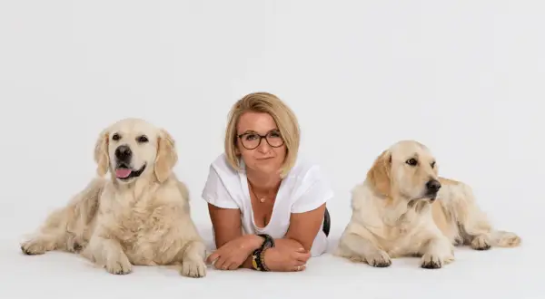 Kobieta w okularach leży między dwoma golden retrieverami na białym tle.