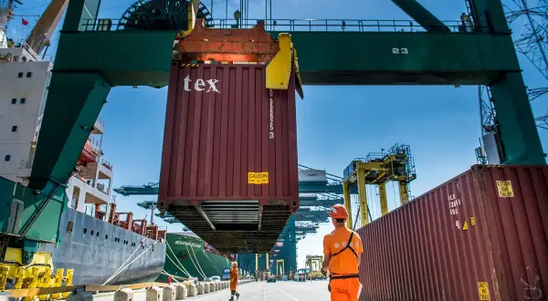 Ein Container wird im Antwerpener Hafen von einem Kran gehoben. Arbeiter in orangefarbener Kleidung überwachen die Aktion.