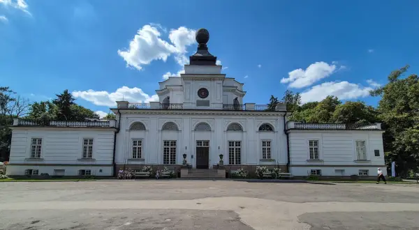 Pałac w Jabłonnie, biała, klasycystyczna budowla z charakterystyczną kopułą, otoczona zielenią, pod błękitnym niebem z chmurami.