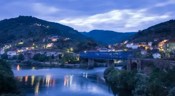Un puente azul cruza un río tranquilo, reflejando las luces de un pueblo en la ladera. La noche cae sobre este paisaje, creando una atmósfera serena.