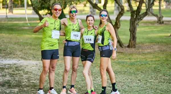 Cuatro corredores sonríen, listos para las carreras populares Zaragoza. Llevan camisetas verdes con dorsales y gafas de sol.