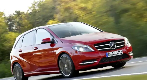 Czerwony Mercedes B Klasa pędzi po drodze, otoczony zielenią drzew.