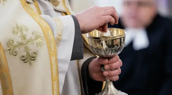 Dłoń księdza w białej szacie z haftem podaje hostię do kielicha. Komunia bez spowiedzi, symbol wiary.