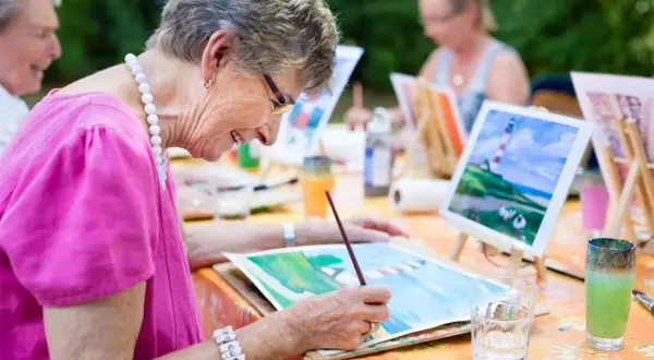 Des personnes âgées participent à des activités manuelles en peignant en plein air. Une dame souriante, vêtue d'un haut rose, est concentrée sur sa toile.