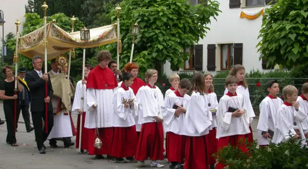 Prozession am Corpus Christi Feiertag mit Ministranten in weißen und roten Gewändern, die eine Monstranz tragen.