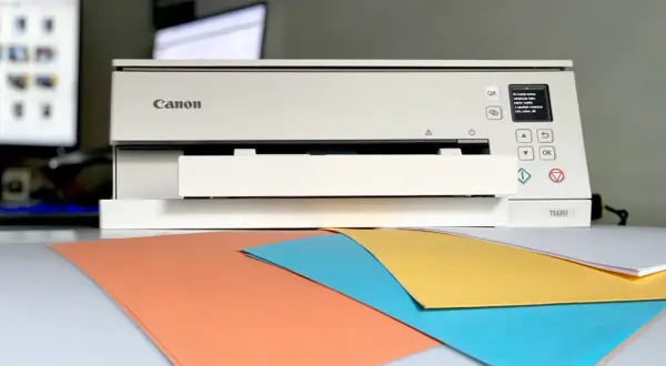 Drukarka Canon TS6351 wyświetla komunikat o błędzie, sugerując, dlaczego z drukarki wychodzą puste kartki. Na biurku kolorowe papiery.