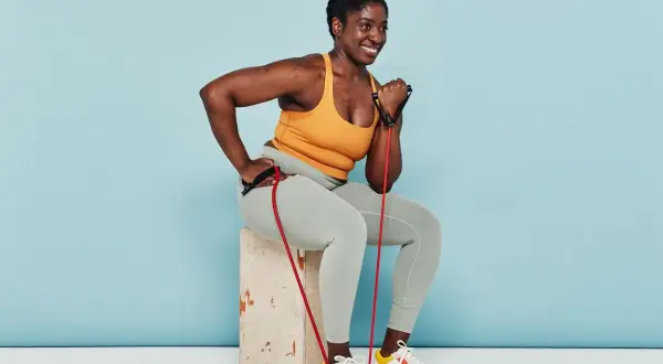 Woman smiles while performing arm strength exercises with a resistance band, seated on a box.